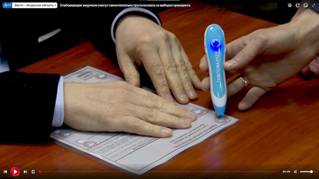 Скриншот из видеосюжета. На нём изображён бюллетень для голосования, на котором лежит рука человека. Рядом другая рука протягивает тифломаркер.