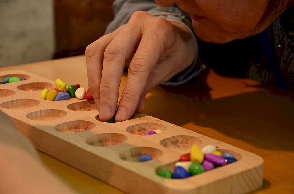 На фотографии игра Каллах. деревянное поле с углублениями, в которые нужно класть разноцветные маленькие камни.. Пальцы человека касаютсяв одного из углублений