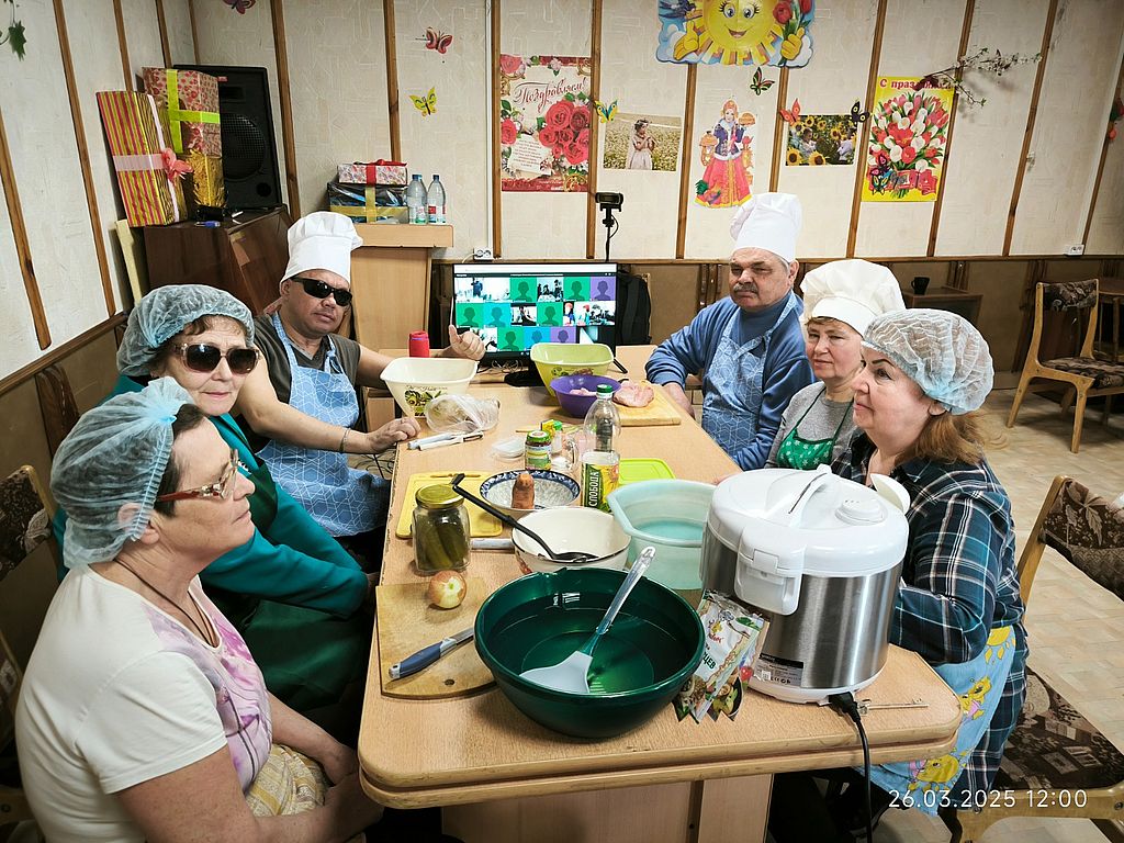 участники онлайн урока сидят за столом, на котором стоит монитор с трансляцией, мультиварка и лежат продукты для приготовления блюда