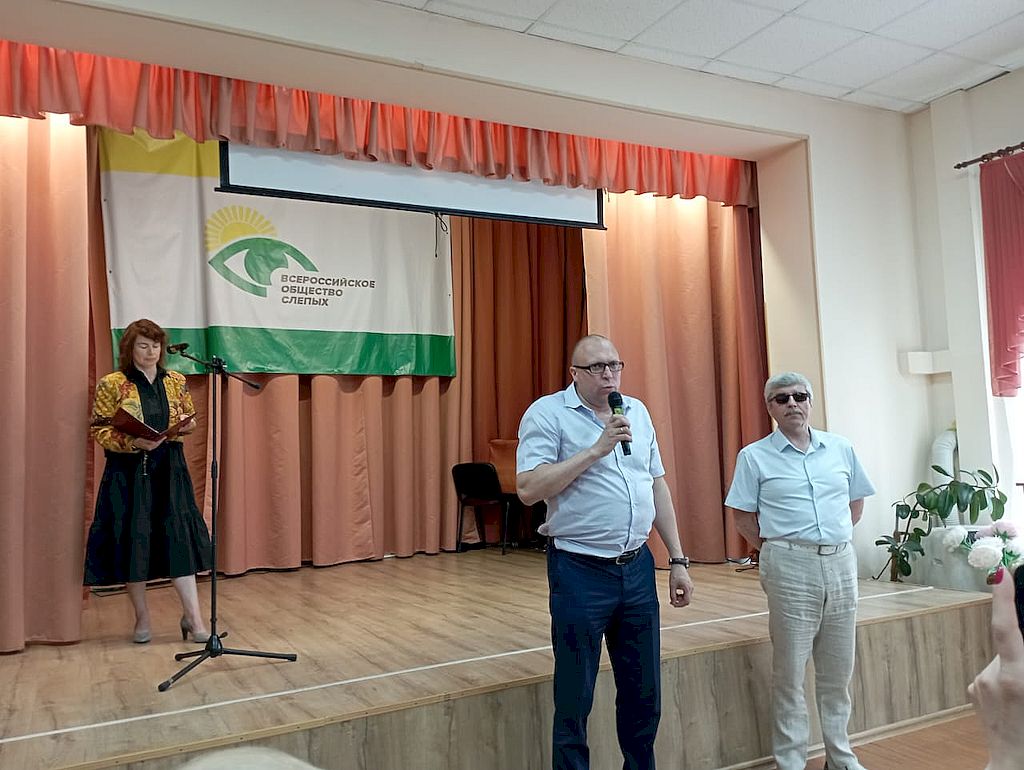 На сцене стоит щенщина, около сцены стоят двое мужчин - один из них с микрофоном. На заднике сцена флаг Всероссийского общества слепых