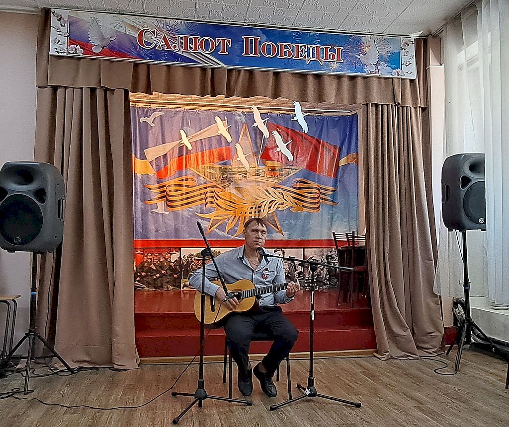 на сцене на стуле сидит мужчина с гитарой. На заднике надпись "Салют Победы", ниже композиция из флага России, георгиевской ленты, белых птиц и фотографии военных