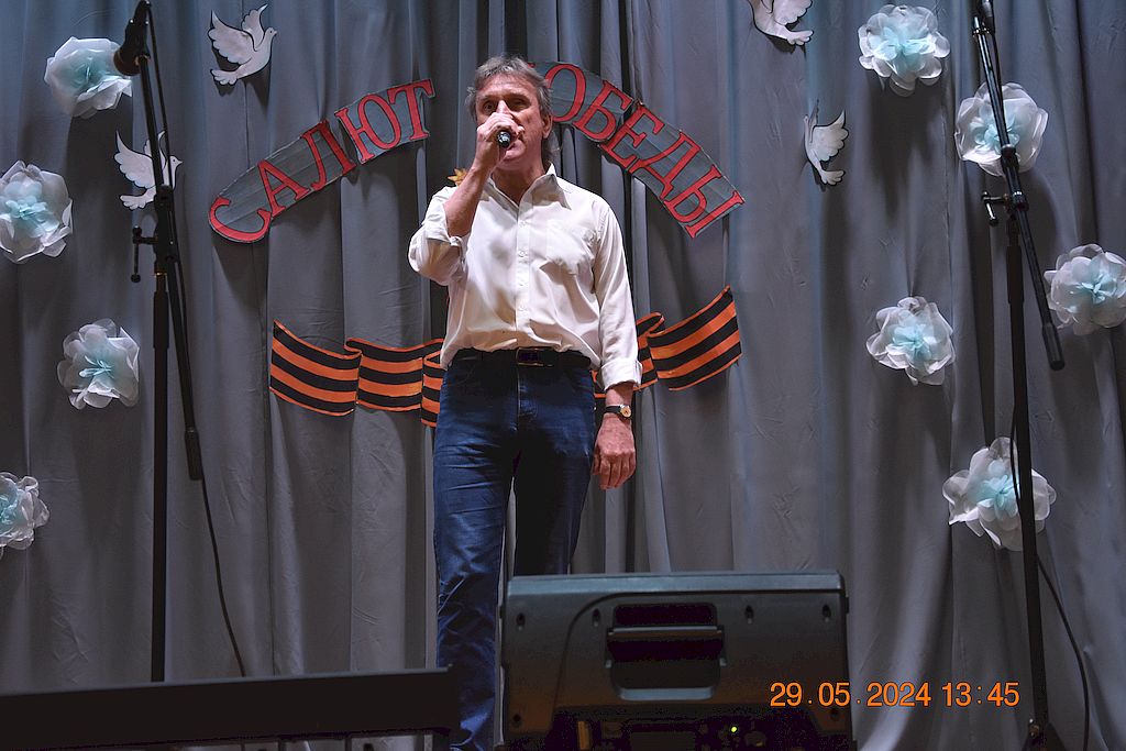 На сцене с микрофоном мужчина в белой рубашке и синих джинсах