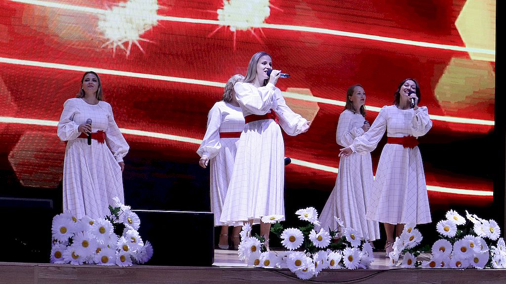 На сцене 5 девушек в белых платьях. Платья перетянуты красными поясами. У ног исполнительниц лежат букеты из ромашек.