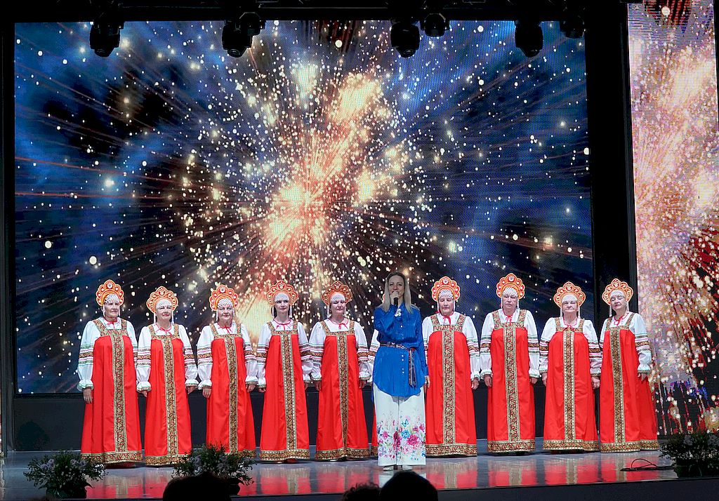 На сцене ансамбль в русских народных костюмах. Перед ними солистка в светлых брюках и синей блузке.