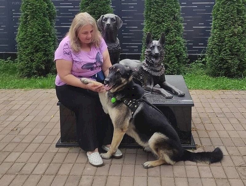 Женщина в чёрных брюках и розовой футболке сидит на краешке памятника собакам-проводникам, который находится в РШПСП ВОС в Купавне.Рядом с ней сидит восточно-европейская овчарка и протягивает женщине лапу.