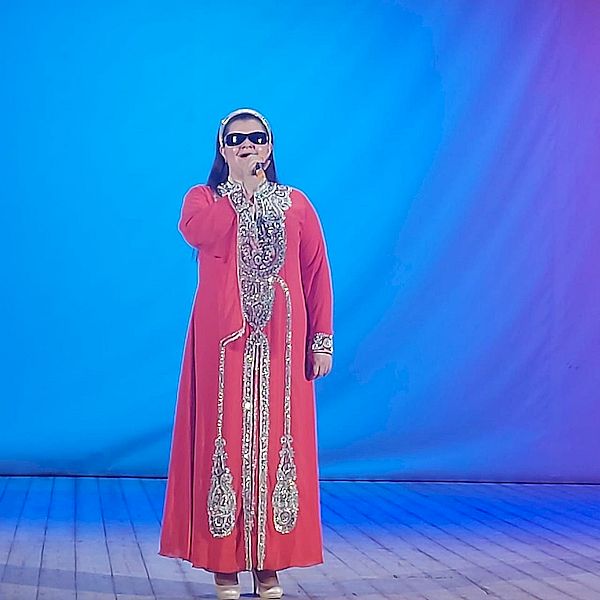 Анастасия Олейникова на сцене с микрофоном в руке. На ней красное длинное платье с серебряными узорами.
