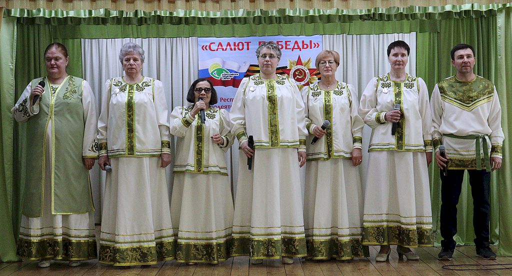 Участники фестиваля в национальных нарядах. Шесть женщин и мужчина. Они одеты в в светлые костюмы с зелёной оторочкой.