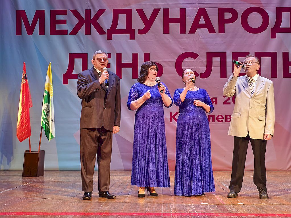 На сцене с микрофонами в руках выступает вокальный квартет «Подолье» Подольской местной организации ВОС. В центре – две вокалистки в синих платьях. Слева вокалист в сером костюме, справа – вокалист в черных брюках и бежевом пиджаке. На заднем плане на флагштоках флаги Московской области и Московской областной организации ВОС