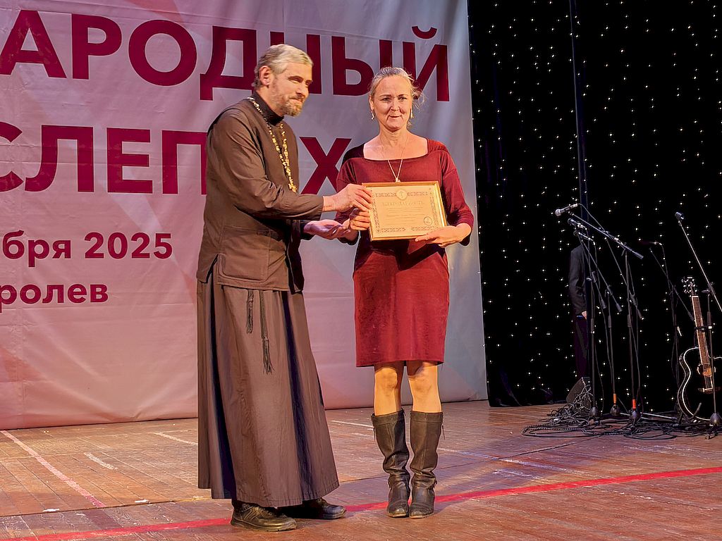 На сцене – иерей Глеб Козлов в церковном одеянии, ответственный по благотворительности и социальному служению Королёвского благочиния. Он вручает председателю Королёвской местной организации ВОС Лилии Богдановой Благодарность Межъепархиального отдела по благотворительности и социальному служению Московской митрополии. Лилия Богданова в платье бордового цвета и черных сапогах. 