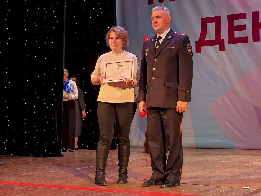 На сцене – начальник отдела пропаганды безопасности дорожного движения Управления Госавтоинспекции Московской области Александр Жубрилов в форме ГАИ. Слева руководитель клуба «Видящие сердцем» Комплексного центра социального обслуживания и реабилитации «Серпуховский» Светлана Смоленцева в белом джемпере, темно-серых брюках и черных сапогах. В руках она держит Грамоту Управления государственной инспекции безопасности дорожного движения Главного управления МВД России по Московской области.