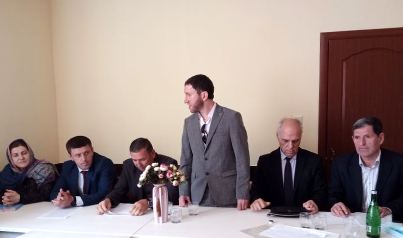 За белым столом шесть человек. слева направо: женщина в платке, пятеро мужчин в костюмах и галстуках. Мужчина посередине стоит и говорит