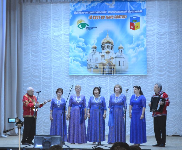 пять женщин в синих платьях поют, по бокам мужчины в красных рубахах играют - один на балалайке, другой на баяне