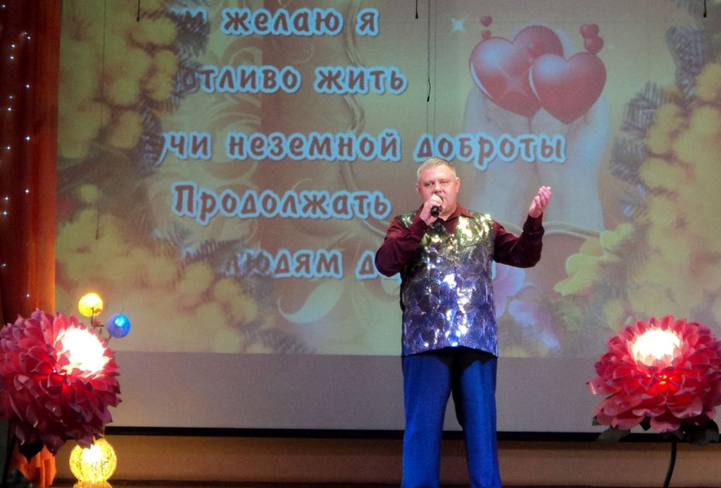 На сцене взрослый мужчина с микрофоном в руке. На нём синие брюки, бордовая рубашка с длинным рукавом и серебристая блестящая жилетка.