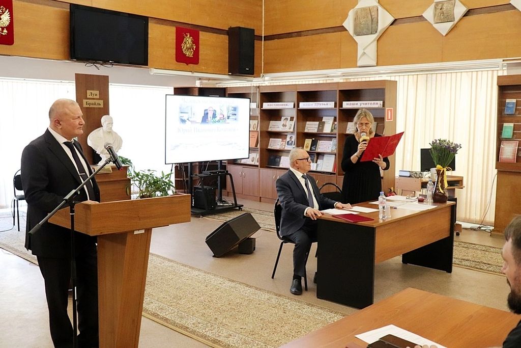 Вице-президент ВОС Э. К. Тедеев стоит за трибуной с микрофоном, справа от него за столом сидит Ю. И. Кочетков , справа от которого стоит взрослая женщина в очках  с микрофоном и красной папкой в руках.