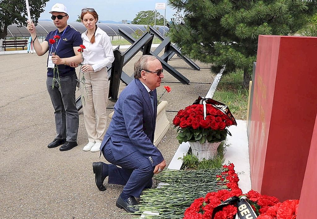 Президент ВОС возлагает цветы к мемориалу. Он стоит на одном колене в синем классическом костюме. На заднем фоне девушка и молодой человек - в руках у них по две гвоздики