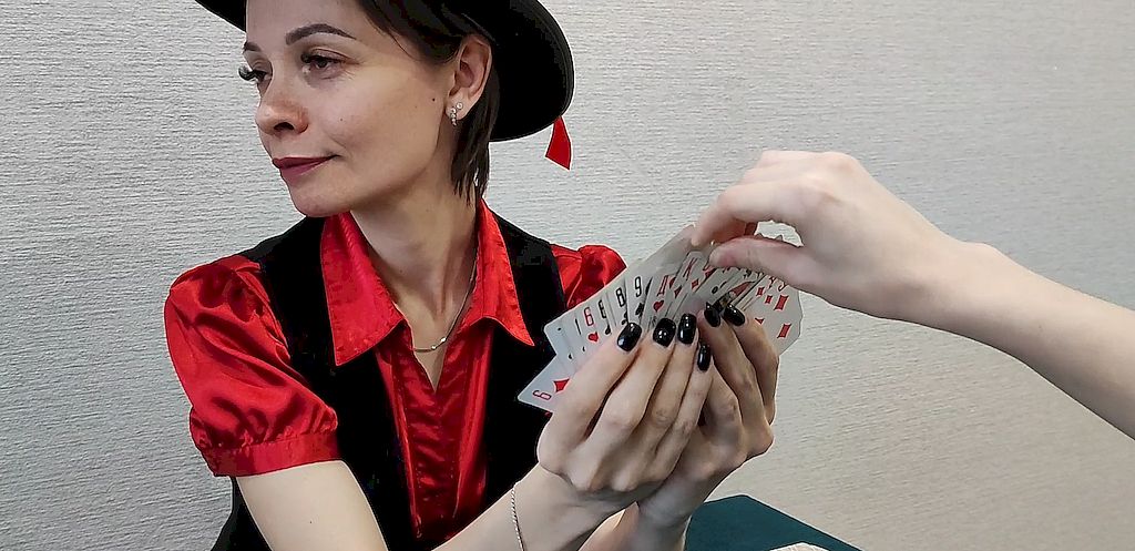 На фотографии девушка в красной рубашке, чёрном сарафане и чёрной шляпе. В руках у неё колода карт, обращённая мастями от неё. Чья-то рука тянется  и забирает карту из колоды