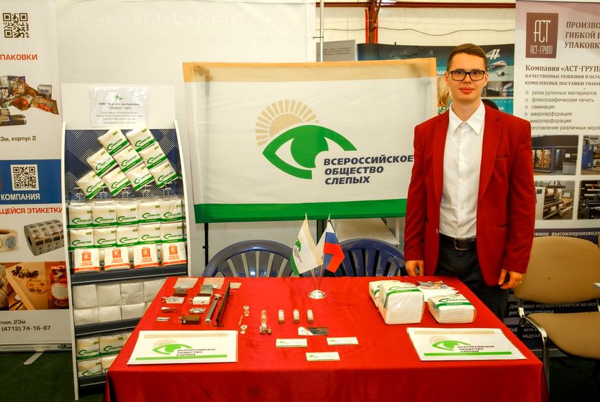 выставка. тизкур представляет свою продукцию. слева стенд с образцами упаковок салфеток. посередине стол с красной скатерью, на нем флажки вос и рф и образцы продукции. за столом баннер с логотипом вос. справа зрячий молодой человек в очках, белой рубашке и красном пиджаке