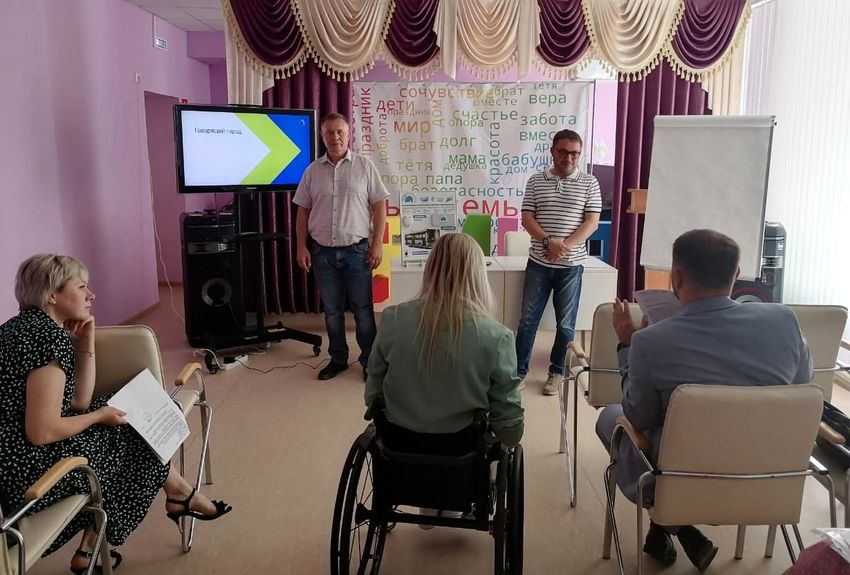 фрагмент совещания. на первом плане сидят три человека, к нам спиной: женщина боком - короткие светлые волосы, черное летнее платье, в руке документ, посередине девушка с длинными светлыми волосами, светло-зеленом костюме на инвалидной коляске, справа мужчина в костюме. на заднем плане - к нам лицом - два мужчины. телевизор - на экране надпись 