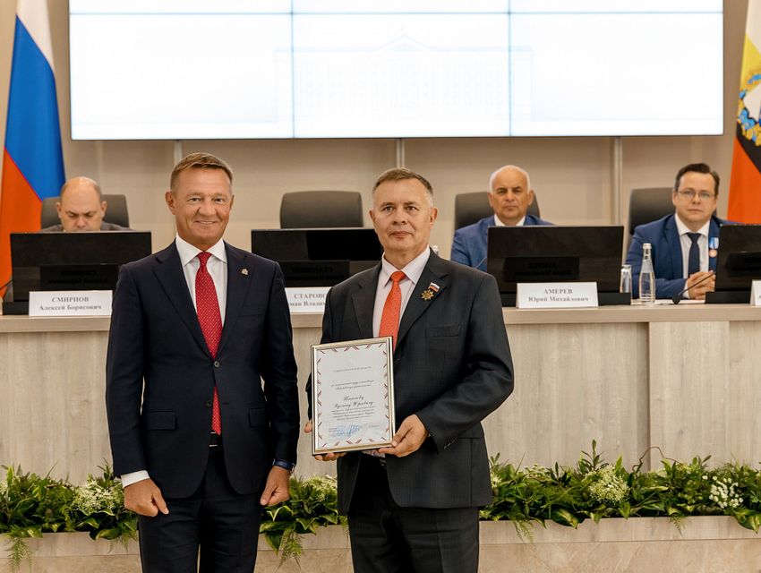 2. Белый зал. На переднем плане губернатор Курской области и председатель Курской региональной организации ВОС. Смотрят прямо в кадр. У председателя в руках плакетка с Благодарностью губернатора. За ними на возвышении белый длинный стол. По бокам флаги, слева РФ, справа Курской области. За столом сидят трое взрослых мужчин в костюмах.