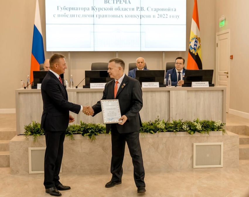Белый зал. На переднем плане слева губернатор, он жмёт руку справа стоящему председателю Курской региональной организации. У председателя в руках плакетка с Благодарностью губернатора. За ними на возвышении белый длинный стол. По бокам флаги, слева РФ, справа Курской области. За столом сидят трое взрослых мужчин в костюмах, над ними экран с надписью: Встреча губернатора Курской области Старовойта Р. В. с победителями грантовых конкурсов в 2022 году.