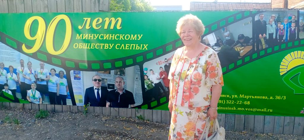 В. И. Прудкова на фоне горизонтально растянутого на заборе баннера. На баннере изображены фотографии  с членами МО ВОС, в том числе фото председателя Минусинской МО ВОС Ф. Ф. Усынина с президентом ВОС В. В. Сипкиным. Фотографии соединены между собой и выглядят как киноплёнка. сверху и снизу от фотографий на зелёном фоне расположены надписи жёлтыми буквами . Сверху: 