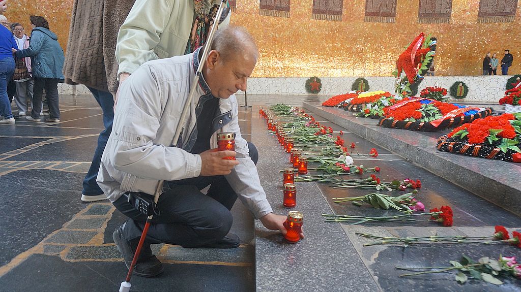 Мужчина с белой тростью ставит зажжённую свечу в банке на постамент в зале Пантеона Славы
