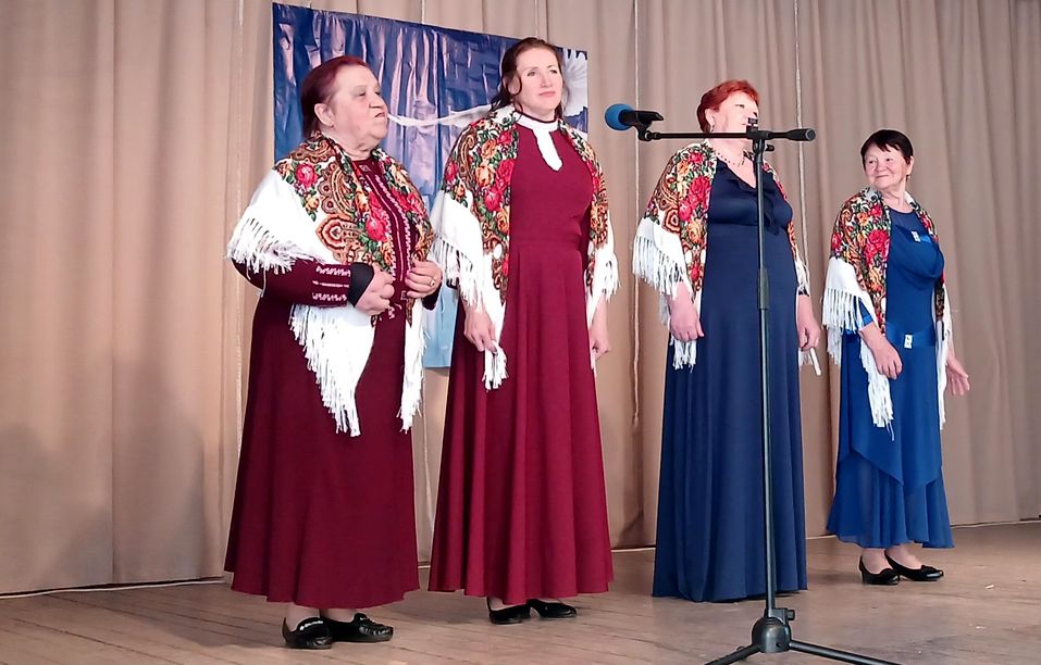 На сцене четыре женщины в сарафанах синего и бордового цвета, с платками на плечах поют у микрофона.