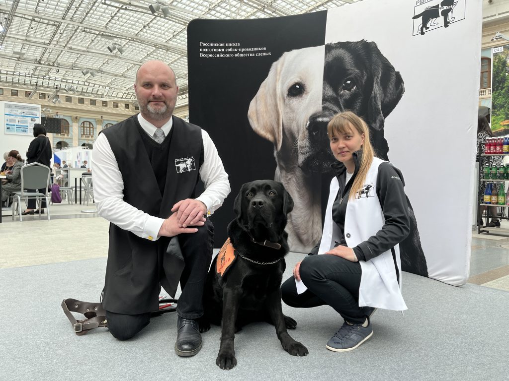 директор РШПСП ВОС Артём Астанин и методист Школы с чёрным лабрадором на фоне баннера Российской школы подготовки собак-проводников