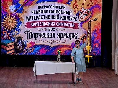 на сцене стоит женщина. рядом с ней стол, на котором стоит ваза с цветами