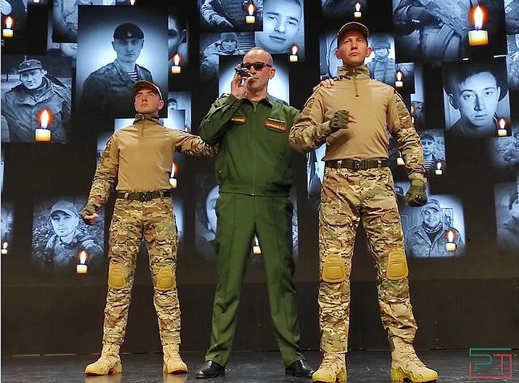 На сцене - 3 человека в форме, один из них -  в тёмных очках