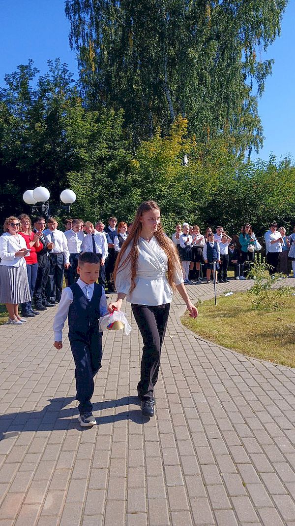 Первый звонок. Старшеклассница ведёт за руку первоклассника. У неё в руке золотистый колокольчик.